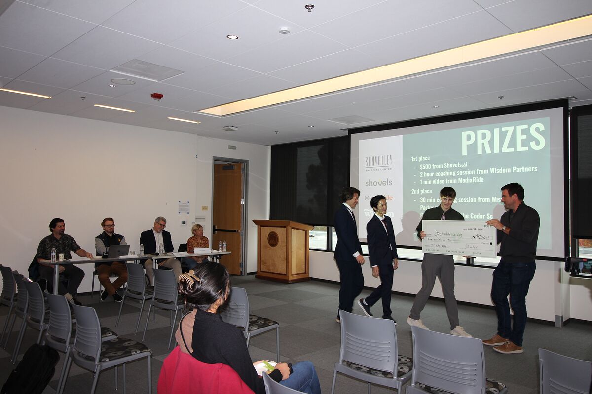 Members of the winning team walk an oversized check toward the front of the room while the judges' table looks on from the left.